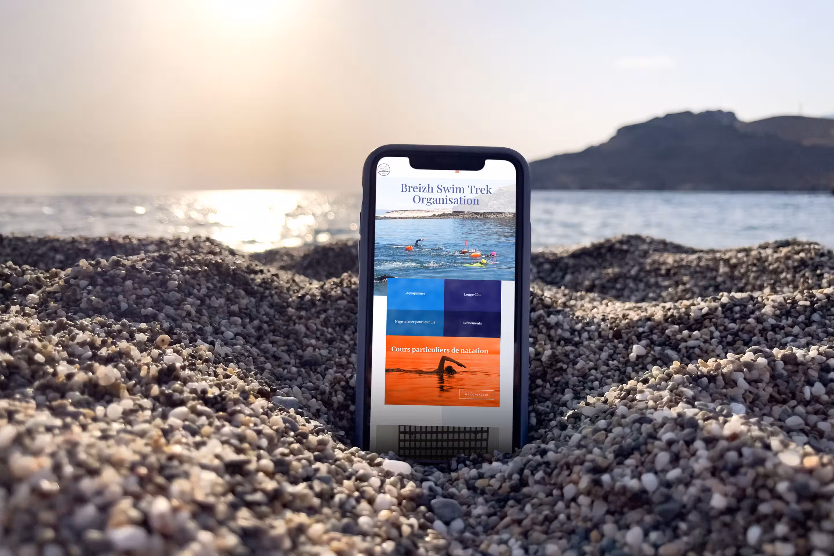Site internet réalisé pour une activité de nage en eau libre, affiché sur smartphone posé sur des galets en bord de mer, exemple de réalisation dans le Morbihan